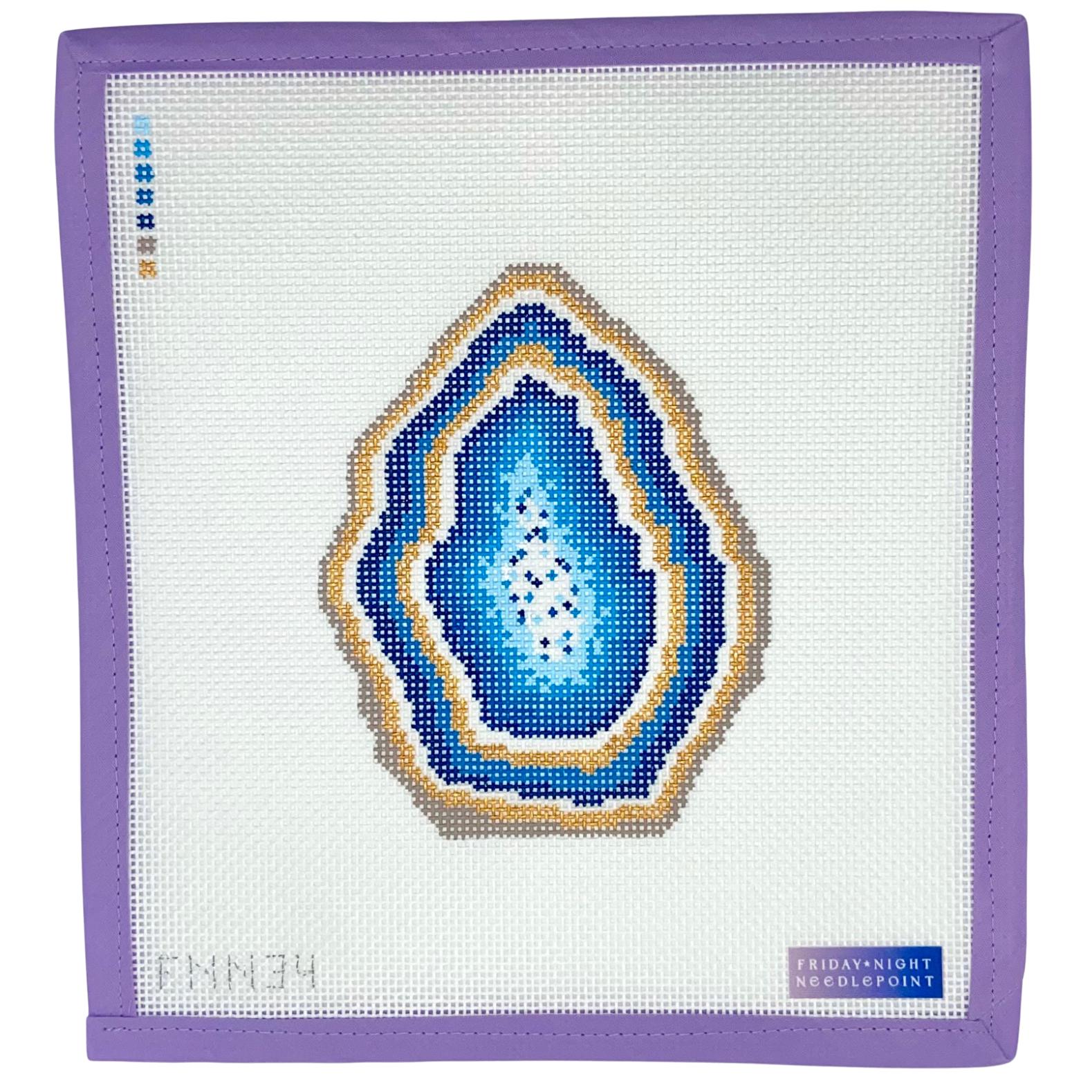 Needlepoint canvas with a blue and gold geode design on a white fabric background, framed by a purple border.