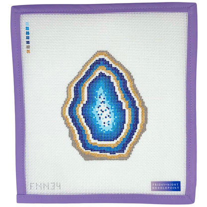 Needlepoint canvas with a blue and gold geode design on a white fabric background, framed by a purple border.